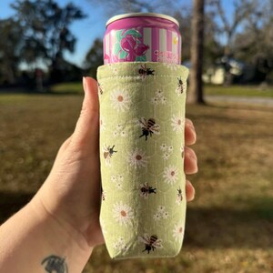 May include: A green fabric can cooler with a pattern of white daisies and black bees, holding a pink and white striped can. The can has a floral design and the word "conto" printed on it.