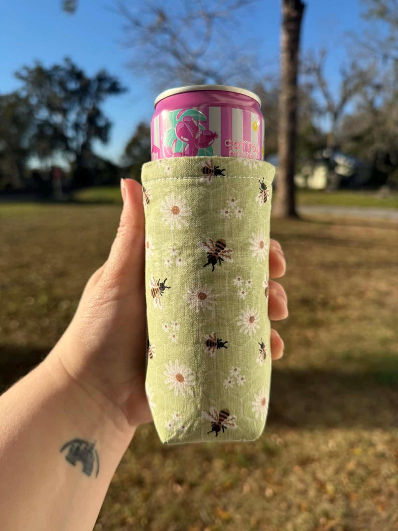 May include: A green fabric can cooler with a pattern of white daisies and black bees, holding a pink and white striped can. The can has a floral design and the word "conto" printed on it.