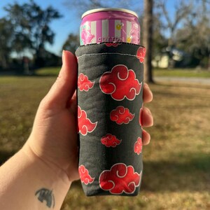 May include: A black cotton can cooler featuring a pattern of red clouds. The cooler holds a pink and white striped can with the word "Cotton" printed on it. The cooler is held in a hand.