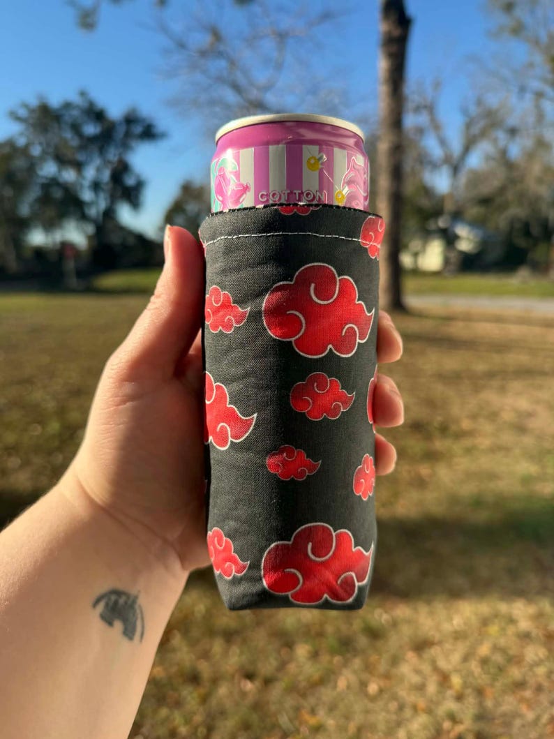 May include: A black cotton can cooler featuring a pattern of red clouds. The cooler holds a pink and white striped can with the word "Cotton" printed on it. The cooler is held in a hand.