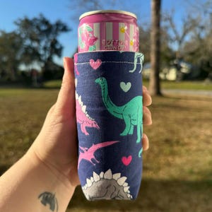 May include: A navy blue can cooler featuring a pattern of pink and green dinosaurs and hearts. A pink and white can of Cotton Candy soda is inside the cooler. The cooler is held in a hand against a background of green grass and a blue sky.
