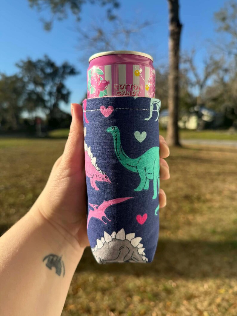 May include: A navy blue can cooler featuring a pattern of pink and green dinosaurs and hearts. A pink and white can of Cotton Candy soda is inside the cooler. The cooler is held in a hand against a background of green grass and a blue sky.