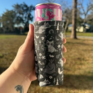 May include: A black and white fabric can cooler featuring a woodland pattern with raccoons, trees, and moons. A pink and white can is inside the cooler. The cooler is held up against a blurred outdoor background.