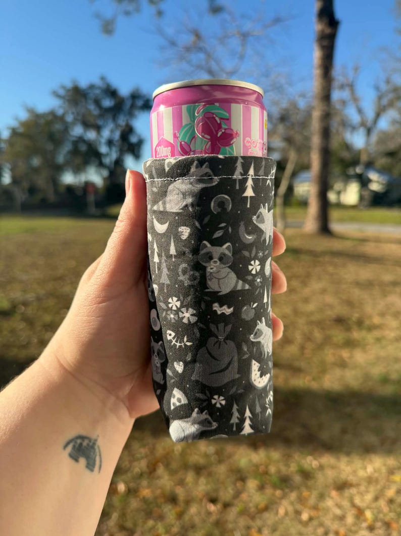 May include: A black and white fabric can cooler featuring a woodland pattern with raccoons, trees, and moons. A pink and white can is inside the cooler. The cooler is held up against a blurred outdoor background.