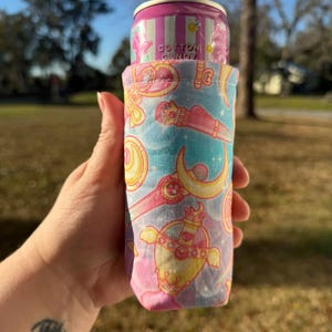 May include: A pink and blue fabric can cooler featuring a repeating pattern of hearts, moons, and other decorative elements. The cooler holds a cotton candy soda can. The can cooler is held by a person.