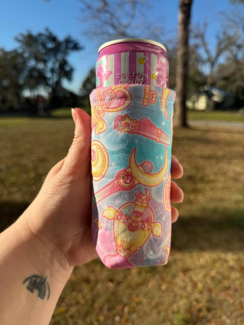 May include: A pink and blue fabric can cooler featuring a repeating pattern of hearts, moons, and other decorative elements. The cooler holds a cotton candy soda can. The can cooler is held by a person.