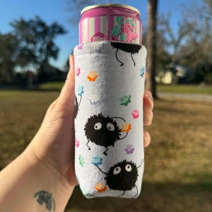 May include: A light blue can cooler featuring black soot sprites and colorful flower accents. The cooler holds a pink and white striped can with a floral design. The can cooler is designed to keep beverages cold.