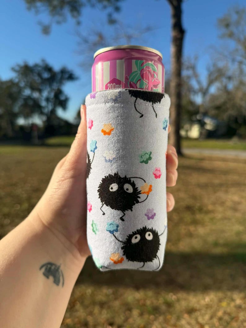 May include: A light blue can cooler featuring black soot sprites and colorful flower accents. The cooler holds a pink and white striped can with a floral design. The can cooler is designed to keep beverages cold.