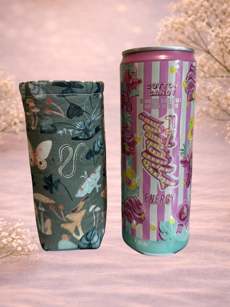 Op de afbeelding: Een grijze stoffen drankhoes met een grillig patroon van paddenstoelen, vlinders en andere natuurlijke elementen staat naast een energiedrankblikje met suikersmaak. Het blikje is roze en wit gestreept met het woord "Energy".