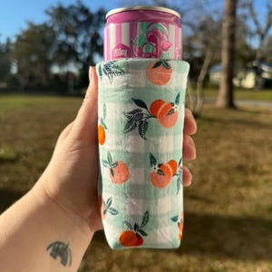 May include: A fabric can cooler with a light green and white checkered pattern, adorned with orange peach illustrations. A pink and white striped can with the text "200mg" is inside the cooler. The cooler is held outdoors.