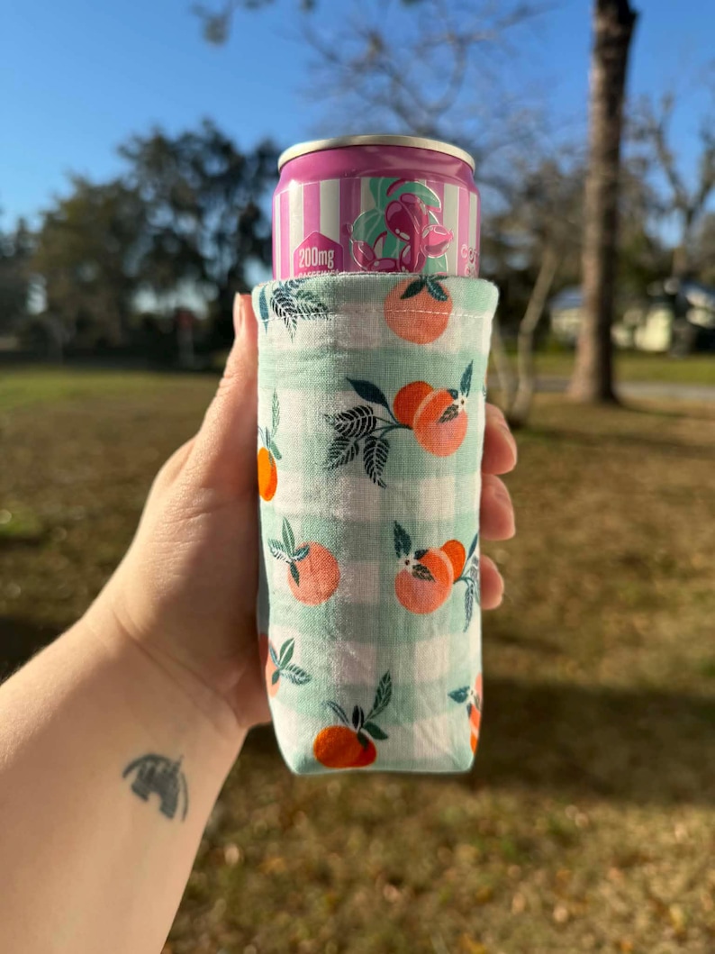 May include: A fabric can cooler with a light green and white checkered pattern, adorned with orange peach illustrations. A pink and white striped can with the text "200mg" is inside the cooler. The cooler is held outdoors.