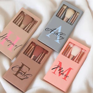 Könnte beinhalten: Vier rechteckige Make-up-Pinsel-Etuis in Beige, Grau, Braun und Pfirsich, jeweils mit einem Sichtfenster, das die Pinsel im Inneren zeigt. Die Etuis tragen personalisierte Namen in Schreibschrift über großen Großbuchstaben: Hacquel, Ashley, Emily und Marc.