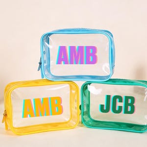 Op de afbeelding: Drie transparante cosmetische tassen met gekleurde randen en gepersonaliseerde initialen. De tassen zijn blauw, geel en groen, elk met een andere set initialen in een levendig 3D-effect. Make-up producten zijn zichtbaar op de achtergrond.