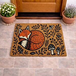A decorative door mat with a fox curled up and surrounded by leaves and mushrooms, placed on a tiled floor near a wooden door.