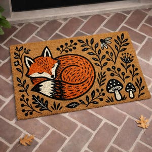 A decorative door mat with a fox curled up and surrounded by leaves, mushrooms, and flowers.
