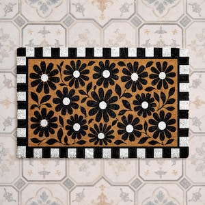 May include: A rectangular doormat with a natural tan background featuring a repeating pattern of black flowers with white centers. The mat is framed by a bold border consisting of alternating black and white squares, creating a high-contrast design.