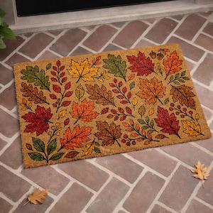 May include: A rectangular doormat with a natural coir background. The mat features a repeating pattern of colorful autumn leaves in shades of red, orange, yellow, and green. The leaves are arranged in a decorative design.