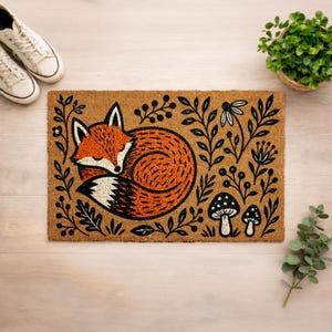 a decorative door mat with a fox illustration and various natural elements such as mushrooms and leaves. The mat is placed on a wooden surface, with a pair of white sneakers and a potted plant nearby.
