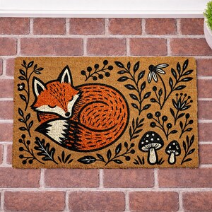 A decorative door mat with a fox curled up and surrounded by various plants and mushrooms.