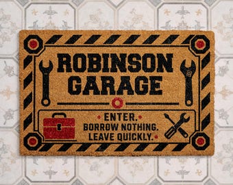 May include: A rectangular tan coir doormat with a black and red border featuring a wrench, a toolbox, and a screwdriver. The text reads: ROBINSON GARAGE. ENTER. BORROW NOTHING. LEAVE QUICKLY. The design includes black diagonal stripes and red bolt accents.