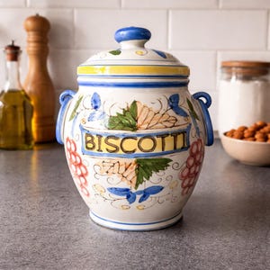May include: A white ceramic biscotti jar with a blue and yellow rim, blue handles, and a blue knob on the lid. The jar is decorated with floral and grape designs and the word "BISCOTTI".