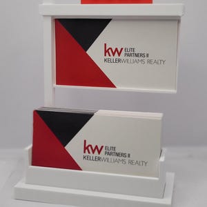 May include: A white real estate sign with a red "SOLD" sign on top. The sign features the "kw ELITE PARTNERS II KELLERWILLIAMS REALTY" logo. Business cards with the same logo are in a holder.