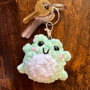 May include: A plush, light green frog keychain with a white belly and pink cheeks. The frog has black eyes and a smiling mouth. The keychain is attached to a silver clasp and a ring holding two keys.