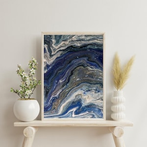 Könnte beinhalten: Ein rechteckiger Wandkunstdruck mit einem abstrakten, flüssigen Acryl-Gießdesign mit wirbelnden Mustern in tiefem Marineblau, Königsblau, Metallic-Gold und Weißtönen, eingefasst in einen hellen Holzrahmen mit einem schlichten, sauberen Rand.