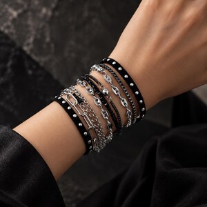 May include: A multi-strand bracelet set featuring a black band with silver studs, a silver-toned paperclip chain, a fine silver chain, a black beaded strand, and a silver-toned mariner link chain, all worn together as a layered wrist accessory.