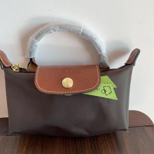 May include: A brown handbag with a leather flap and handle. The bag has a gold-colored clasp and a small, round gold-colored button on the flap. A green tag is tucked inside the bag.