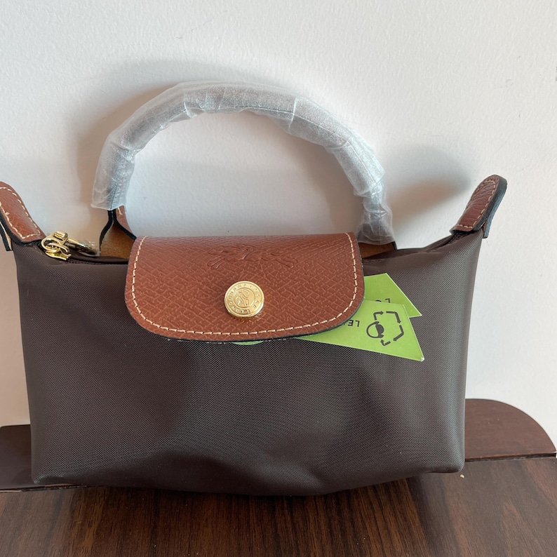 May include: A brown handbag with a leather flap and handle. The bag has a gold-colored clasp and a small, round gold-colored button on the flap. A green tag is tucked inside the bag.