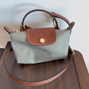 Puede incluir: Un bolso gris con solapa y asa de cuero marrón. El bolso tiene un cierre dorado y una correa marrón desmontable. El bolso está hecho de un material de nailon y tiene un pequeño botón redondo dorado en la solapa.
