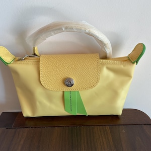 May include: A yellow handbag with a flap closure and a top handle. The bag has a light yellow body with a darker yellow flap and handle. The bag has a silver-colored zipper and a green tag.