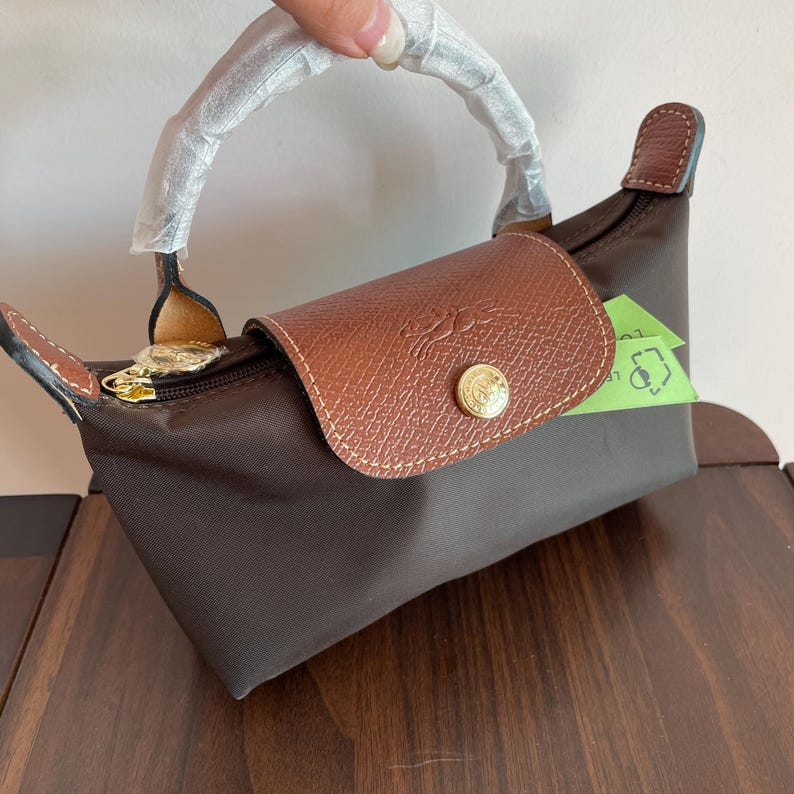 May include: A brown handbag with a leather flap and handle. The bag has a gold zipper and a gold button closure. The leather flap features a logo. The handle is wrapped in white plastic. The bag is made of a nylon material.