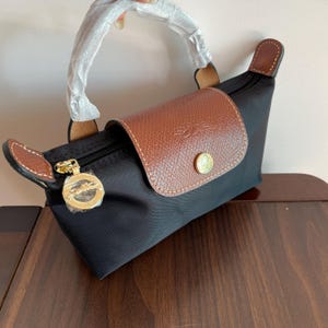 May include: Black handbag with a brown leather flap and handle details. The bag has a gold-toned zip and a round gold emblem. The bag is made of a durable material and has a classic design.