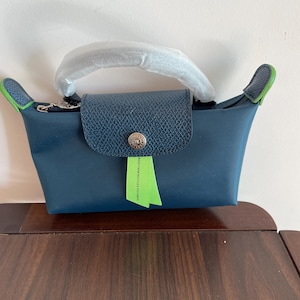 May include: A navy blue handbag with a flap closure and a silver button. The bag has a handle and green accents on the sides. A green tag with white text is attached to the bag.
