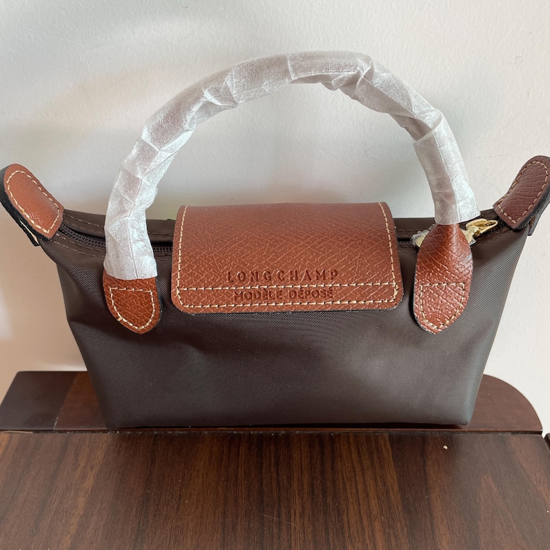 May include: A brown handbag with a brown leather flap and handle. The bag has a zipper closure and brown leather accents. The text "LONGCHAMP MODELE DEPOSE" is visible on the flap. The handle is wrapped in white paper.