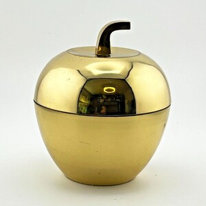 May include: A polished gold-colored metal apple-shaped container with a removable lid. The object features a smooth, reflective surface and a curved stem at the top. The design consists of two main parts that fit together to form a rounded fruit shape.