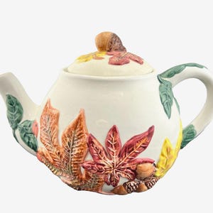 May include: Cream-colored ceramic teapot with a lid and handle. The teapot is decorated with colorful, raised relief autumn leaves in shades of red, orange, and yellow. The lid is topped with a brown acorn.