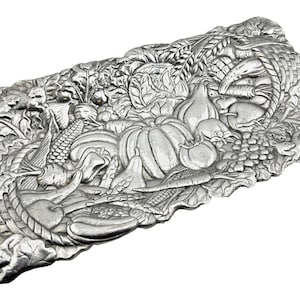 Puede incluir: Una bandeja decorativa rectangular plateada con un diseño en relieve detallado. La bandeja presenta una cornucopia de elementos de la cosecha, incluyendo calabazas, tomates, uvas, maíz y un conejo. Los bordes están intrincadamente esculpidos con hojas y otras formas naturales.