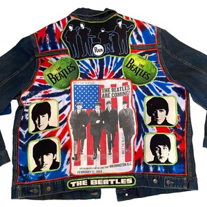 Puede incluir: Una chaqueta de mezclilla azul oscuro con un panel trasero de patrón tie-dye rojo, blanco y azul. Muestra imágenes de The Beatles, incluyendo retratos individuales, una silueta grupal, dos logotipos de manzanas verdes y un póster de Meet The Beatles.