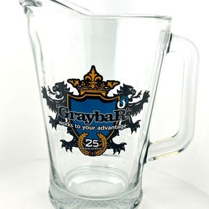 May include: Clear glass pitcher with a handle. The pitcher features a logo with a blue shield, gold crown, and the text "Graybar works to your advantage." Below the logo is a gold emblem that reads "25th Anniversary."