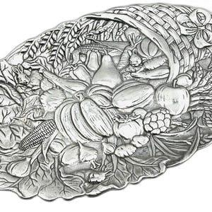Puede incluir: Plato decorativo ovalado de color plateado con un diseño de cornucopia. El plato presenta una cesta tejida que rebosa con representaciones detalladas de varias verduras, frutas y un pequeño conejo. El diseño incluye calabazas, manzanas y maíz.