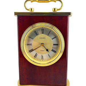 May include: A rectangular, wooden clock with a gold-tone face and accents. The clock face features Roman numerals and the word "Linden". A gold handle sits atop the clock. The clock is approximately 6 inches tall.