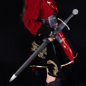 May include: A longsword with a black leather-wrapped scabbard, a silver-toned pommel, and a crossguard. The scabbard features a black leather suspension system with metal buckles and a silver-toned chape at the pointed end of the scabbard.