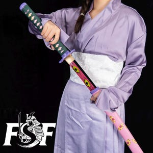 Könnte beinhalten: Dekoratives Katana-Schwert mit rosa und gelbem Blumendesign auf der Scheide. Der Griff ist grün mit rautenförmigen Akzenten. Die Person trägt einen lila Kimono und einen weißen Obi.