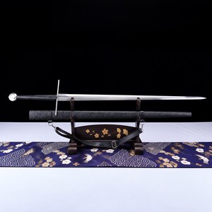 May include: A longsword with a silver-toned blade, a black cord-wrapped grip, and a circular pommel. The sword rests horizontally on a dark wooden stand alongside a black scabbard featuring a black leather carrying strap with a metal buckle.