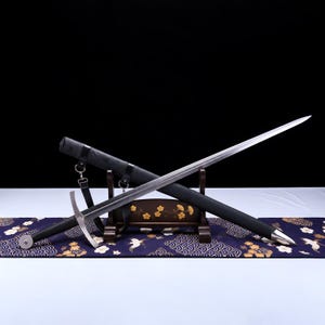 Könnte beinhalten: Ein Langschwert mit silberfarbener Klinge, schwarzem Griff und rundem Knauf, das auf einem dunklen Holzständer ruht. Eine schwarze Schwertscheide mit silberfarbenen Metallbeschlägen und schwarzen Lederriemen liegt diagonal dahinter.