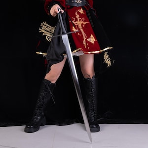May include: A long, double-edged steel sword with a black hilt and a straight crossguard. The blade is silver-toned and tapers to a point. The sword is held vertically, with the tip pointing towards the ground, showcasing its metallic finish.