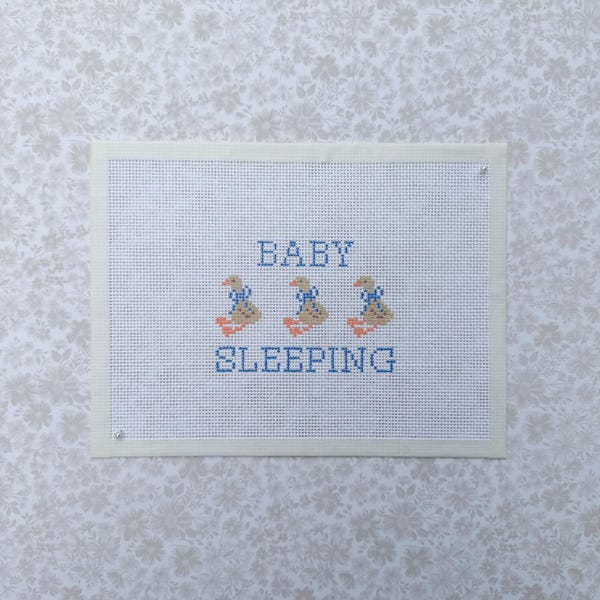 Könnte beinhalten: Ein rechteckiger weißer Aida-Stoff mit einem Kreuzstichmuster. Der Text BABY SLEEPING ist mit blauem Garn gestickt. Zwischen den Wörtern befinden sich drei kleine hellbraune Enten mit orangefarbenen Füßen und blauen Schleifen um den Hals.